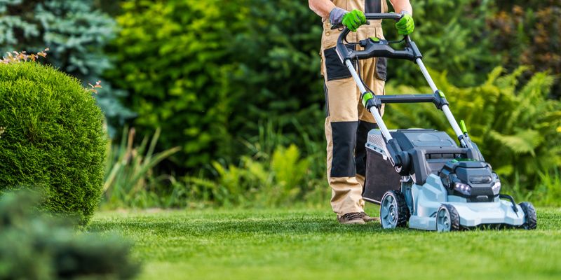 Gardener wearing work gloves, protective work pants and sturdy boots mowing a green lawn with a cordless push mower