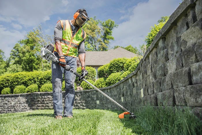 Worker wearing a helmet, face shield and hearing protection trimming grass along a stone wall with a STIHL loop-handle brush cutter